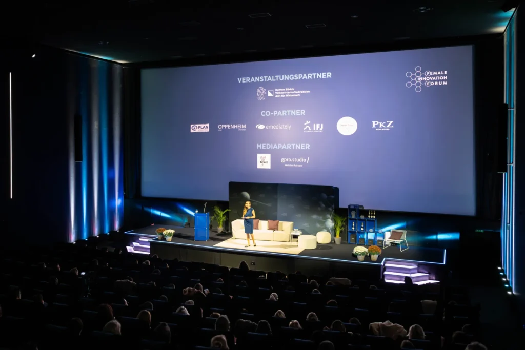 Moderatorin Sunnie Groeneveld auf der Bühne des Female Innovation Forum 2026 vor einer Leinwand mit den Logos der Co-Partner.