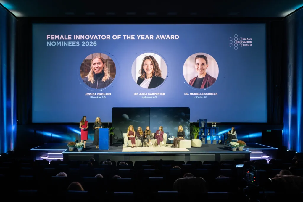 Sandra-Stella Triebl präsentiert auf der Bühne des Female Innovation Forum 2026 die drei Nominierten für den Female Innovator of the Year Award. Auf der Leinwand sind Jessica Droujko, Dr. Julia Carpenter und Dr. Murielle Schreck zu sehen.